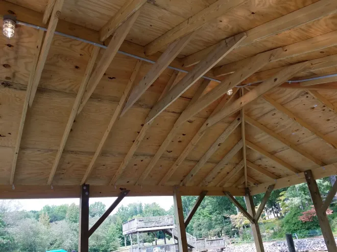 Wooden gazebo interior with exposed rafters and plywood ceiling. Electrical conduit and two lights visible.